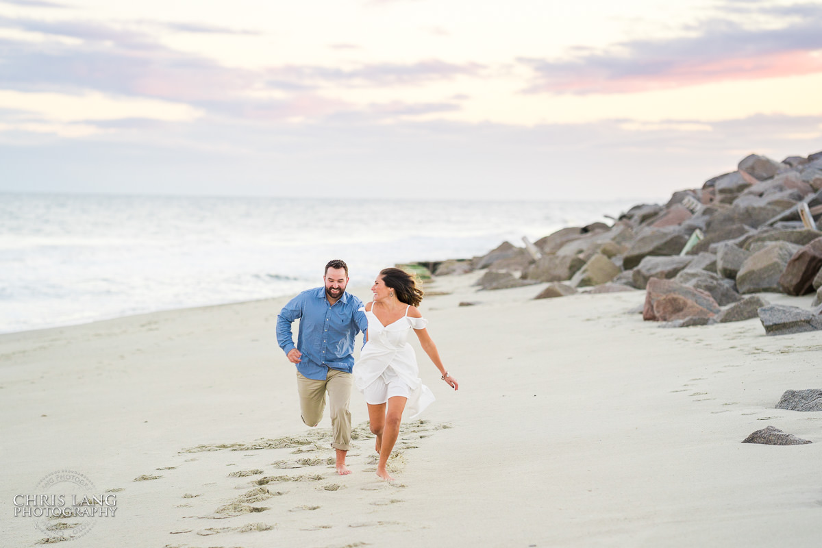 Couple running on the beach - Bright and Airy Engagement photo - Couple wrapped  in Christmas lights on the beach - Fort Fisher North Carolina -  Engagement Photography - Wedding Ideas - Popular engagement photography locations - Lifestyle engagement photo -  Ft Fisher engagement photographers - Engagement session ideas - New trends in engagement photography - Chris Lang Photography - Engagement photos 
